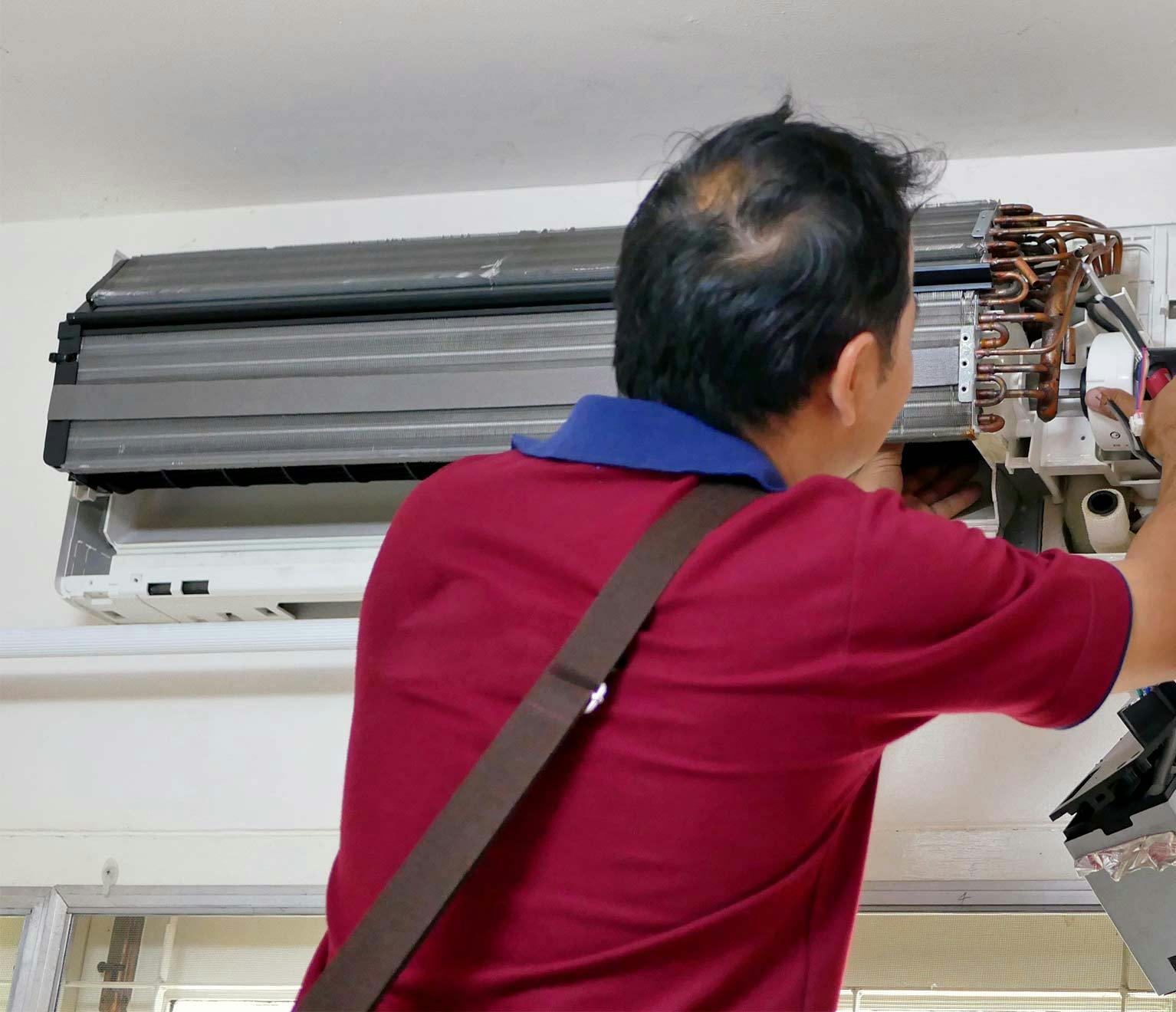 Older man in a red shirt performs repairs on a broken air conditioning unit.